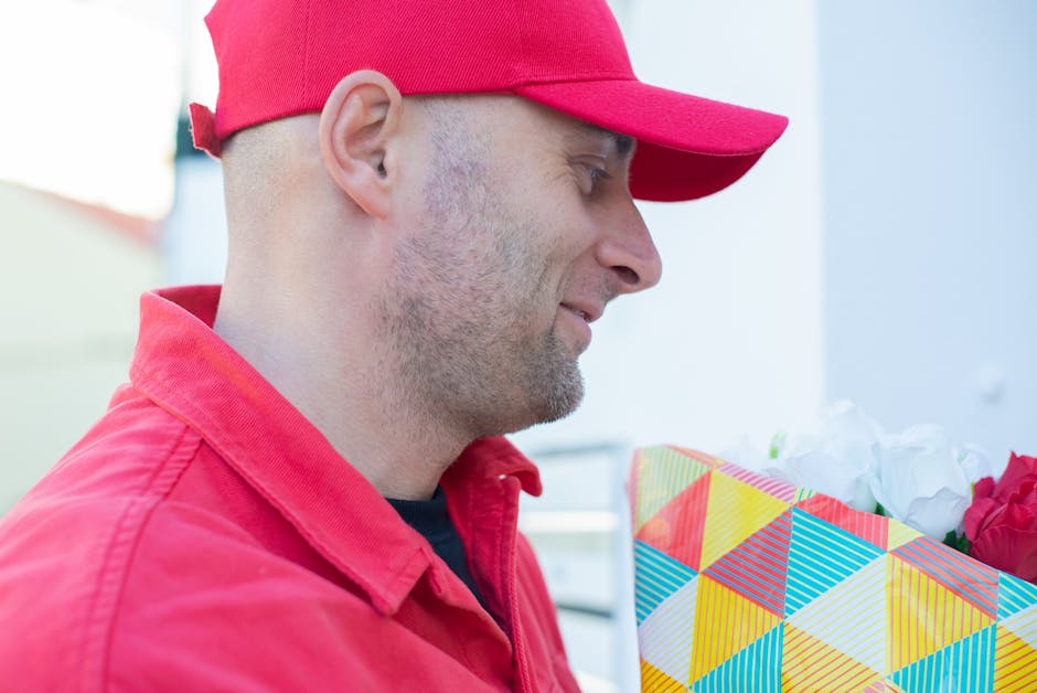 A delivery man wearing a red cap and red jacket holding a vibrant floral gift wrapped in colorful geometric-patterned paper with white tissue paper peeking out, standing outdoors in daylight. The arra