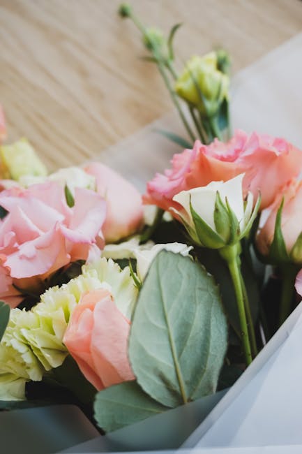 A close-up of a floral bouquet arranged with soft pink and white lisianthus flowers, complemented by fresh green leaves and stems, wrapped in white tissue paper. The bouquet rests on a light wooden su