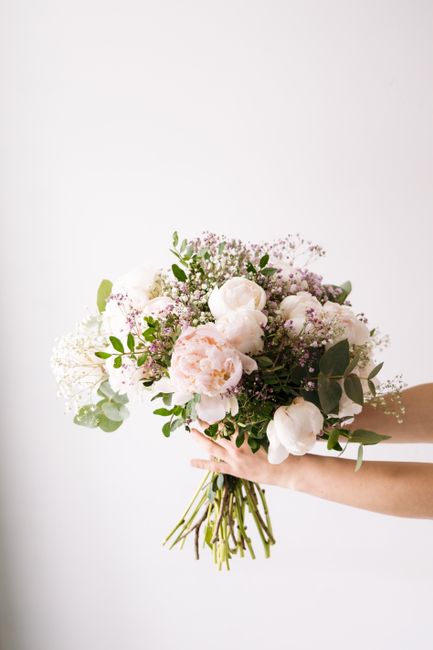 A person holding a fresh floral bouquet featuring large, soft pink and white peonies, complemented by delicate white baby's breath and green foliage, with the stems gathered and wrapped in natural twi