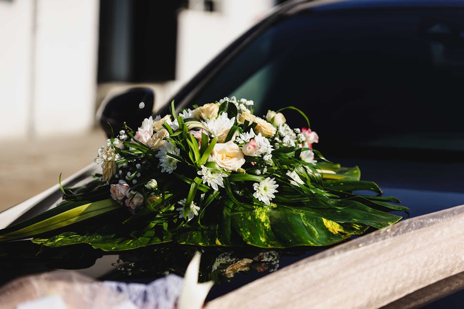 A delicate floral arrangement featuring soft pink and white roses, white daisies, and baby's breath, arranged with lush green foliage on a large green leaf base. The bouquet is placed on the hood of a