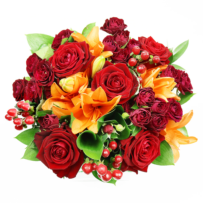 Presented from above on a clean white surface, this perfectly rounded bouquet is a study in warm tonal harmony and thoughtful craftsmanship. Deep red roses, rich and velvety, form the central motif, each bloom opening into layered petals with a soft, tactile quality that suggests timeless devotion. Bright orange Asiatic lilies interrupt the red plane with trumpet-shaped petals and prominent stamens, some blooms fully revealed while others sit in tight, graceful buds that promise unfolding drama over the next days. Tucked into the negative spaces are clusters of small, dark burgundy spray roses whose concentrated, fleshy petals add a lush, layered density and deepen the bouquet's jewel-like palette. Glossy hypericum berries in a vibrant ruby red are sprinkled through the arrangement, offering playful punctuation and a slightly prickly texture against the softer flower faces. Generous swathes of glossy green foliage - ruscus or salal - peek between blooms, giving the composition structure and fresh contrast while keeping the overall silhouette full and rounded. The lighting is soft and even, lifting the sheen on the leaves and the velour of each rose without casting strong shadows, which helps the colours read true and saturated. This ensemble suggests occasions of warmth and celebration: romantic gestures, milestone anniversaries, or a congratulatory gift to accompany a West Norwood dinner invitation. The bouquet feels both opulent and intimate, as if designed by a local hand to brighten a home on Crown Lane or to accompany flowers ordered for a neighbour's celebration.