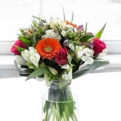 With a balanced palette and dynamic form, this floral arrangement reflects the vibrant yet comforting character of West Norwood living. Creamy peach and vivid orange gerbera daisies cluster at the arrangement's heart, evoking the cheerful tones of summer picnics in Brockwell Park. Deep fuchsia roses, their petals rich and velvety, weave a sense of romance and depth into the bouquet. White alstroemeria and assorted petite white blossoms are scattered artfully throughout, adding a light and fresh texture reminiscent of neighbourhood gardens in bloom. The design finds stability and elegance in the broad green aspidistra leaves that curl gracefully around the base, forming a lush collar alongside the tall, clear glass vase that neatly showcases verdant stems. Gentle, natural light washes across the blooms, reflecting the inviting ambiance of West Norwood's local shops and residential streets. With its gentle fragrance, sophisticated composition, and uplifting palette, this bouquet is the perfect centrepiece for gatherings, celebrations, or thoughtful gifts within this welcoming South London community.
