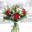 A luxurious red and white bouquet stands proudly in a clear cylindrical glass vase, the kind you might find gracing a sunny dining table in a West Norwood townhouse. Below the waterline, neatly trimmed green stems line up in a tidy cluster, their freshness visible through the transparent glass. Above, velvety deep red roses and a large, ruffled crimson carnation form the passionate heart of the arrangement, their petals unfurling in soft, layered spirals that catch the light. Around these, creamy white alstroemeria with fine speckles and a scattering of white chrysanthemum or daisy-like blooms add a gentle contrast, while several closed white lily buds rise elegantly, promising more beauty in days to come. Wisps of delicate gypsophila, or baby's breath, float between the bolder flowers like tiny clouds, giving the bouquet an airy, romantic softness. Framing everything is lush greenery: broad, glossy dark green leaves are carefully curled and wrapped to create a structured collar at the base, while slimmer eucalyptus-style sprigs and pointed foliage weave upwards, adding layers of texture and varying shades of green. The bouquet sits on a pristine white surface, against a bright, softly blurred background that suggests daylight streaming through a nearby window, much like the calm light along Auckland Hill or near West Norwood station. The overall mood is elegant yet welcoming, a classic colour palette suited to anniversaries, birthdays, Mother's Day, or to bring a warm, heartfelt touch to a local office or living room, radiating affection and quiet celebration.