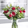 A rich, romantic bouquet of deep crimson red roses, matching ruffled carnations, and crisp white alstroemeria stands proudly in a clear glass vase on a bright white tabletop, bathed in soft natural light from a large window. Each rose shows off velvety petals, some fully open in luxurious spirals, others still in elegant bud form, promising days of unfolding colour. The carnations echo the same intense red, their frilled petals adding fullness and a slightly nostalgic charm, like the classic arrangements often chosen for anniversaries in West Norwood. Nestled between the bold reds, clusters of white Peruvian lilies offer a gentle contrast; a few blooms are partially open, revealing their slender, trumpet-like petals tipped with a faint green, while many buds remain closed, suggesting future beauty. Tiny sprays of white gypsophila scatter like a fine, cloudlike mist throughout the bouquet, softening the composition and catching the diffused daylight. Around the base, broad, glossy green leaves form a supportive collar, giving the display structure and a feeling of lush abundance, while slimmer, lighter green stems weave through, adding movement and depth. The neatly trimmed stems are clearly visible through the transparent vase, submerged in clean water that reflects the care of a local florist who knows how important freshness is for same-day delivery across Norwood Road and nearby streets. Behind the arrangement, the view through the window fades into a softly blurred, almost monochrome scene, so the vibrant reds, whites, and greens take full focus. The overall impression is of elegance, warmth, and celebration, perfectly suited to mark a special occasion, from a romantic gesture to a heartfelt thank you in the West Norwood community.