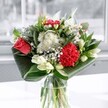 A clear glass vase holds a thoughtfully composed bouquet that feels like a cool breath of air in a calm West Norwood morning. At the heart, a single coral-red rose and a plush crimson carnation glow with velvety depth, their ruffled and layered petals giving the arrangement its confident, modern classic feel. Around them, clusters of tiny daisy-like white blooms and button-shaped chrysanthemums float like soft clouds, their crisp petals adding a sense of purity and gentle joy. Several creamy white lilies sit partly in bud, their long, elegant forms rising slightly above the rest, hinting at blooms still to come and suggesting new beginnings or quiet celebration. Broad, glossy green leaves are carefully folded and curved in wide arcs around the base, wrapping the stems in a sculptural embrace that frames the flowers and emphasises the bouquet's clean, architectural silhouette. The vase stands in a window-lit space, where cool, natural daylight spills across the petals, picking out subtle tones of red, cream, and fresh green, and causing the glass to shimmer lightly. You can almost sense a light, leafy fragrance with a whisper of sweetness, like stepping into a garden after rain along one of the residential streets off Knights Hill. The overall mood is uplifting yet composed, perfect for a joyful occasion, a warm thank-you, or simply brightening a quiet corner of a West Norwood home or office with a touch of refined freshness and heartfelt sentiment.